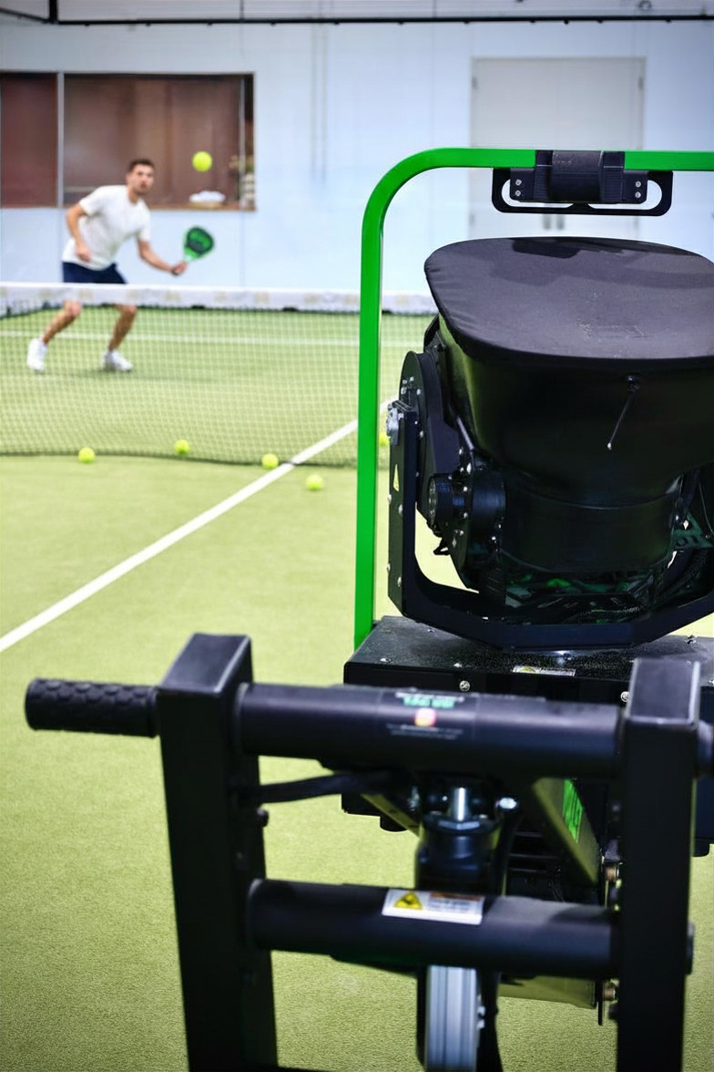 A Volley ball machine on a padel court with a player drilling behind it